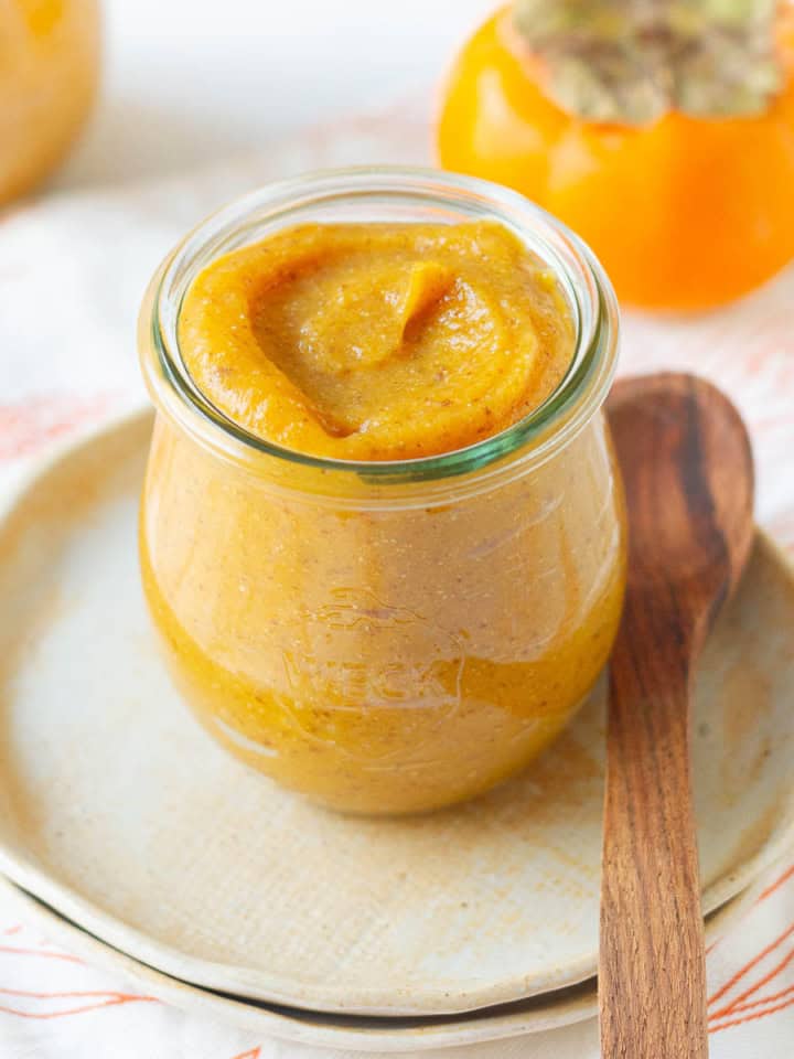 persimmon chia jam in a glass jar with a wooden spoon on a small plate.